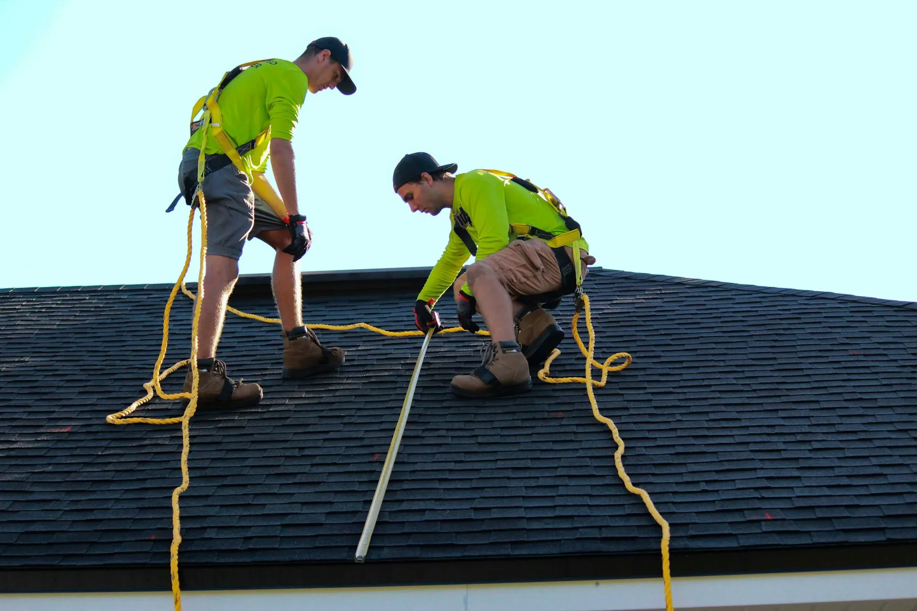 Two roofers in high-visibility shirts working with safety ropes on a dark shingle roof under a pale sky