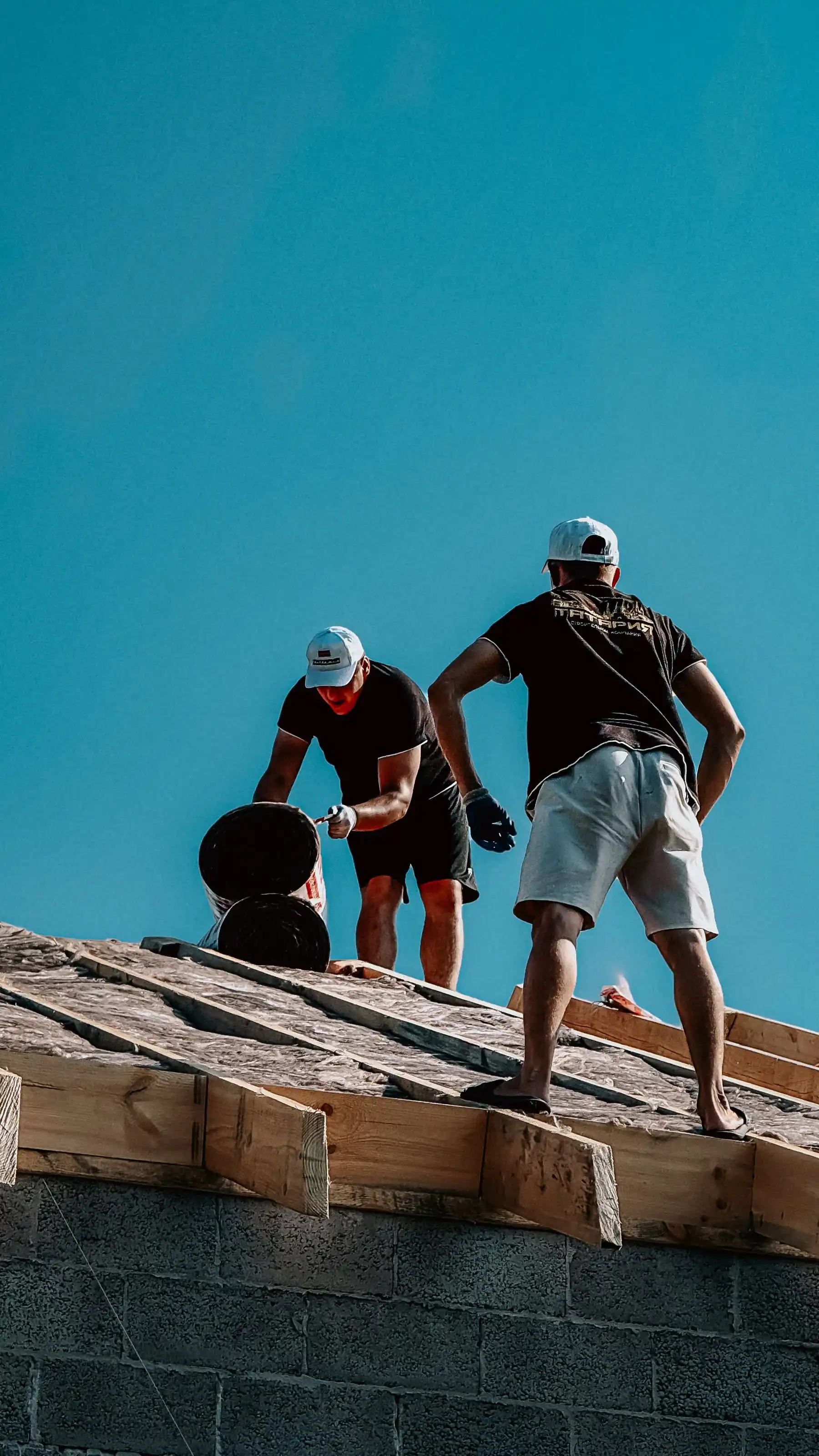 Two roofers working on a roof edge against a bright teal sky