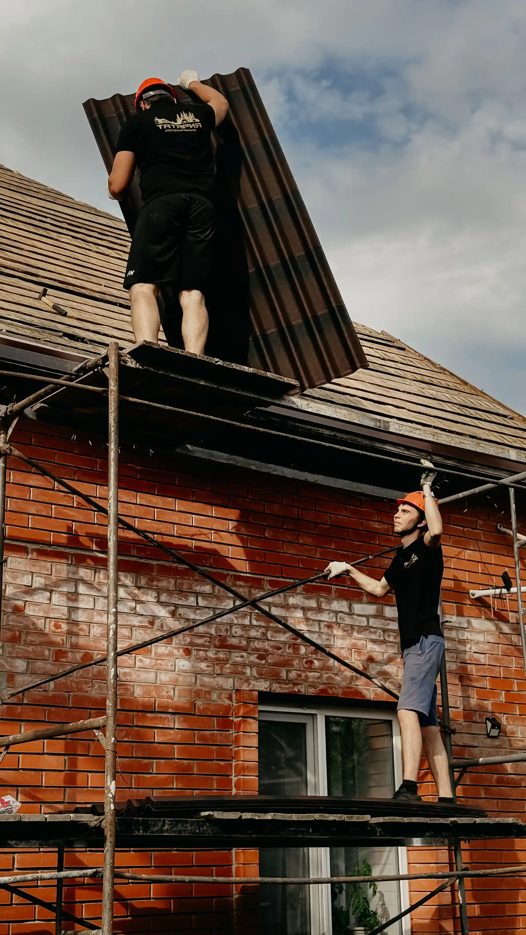 Two builders on scaffolding installing roof panels on a brick building